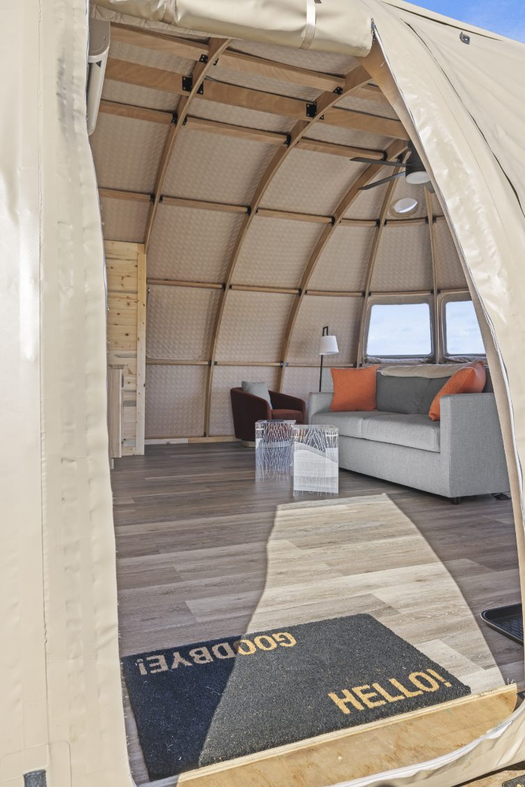 Alt text: "Interior view of a stylish geodesic dome with wooden framework, featuring a cozy seating area with a gray sofa and orange cushions, a modern lamp, and porthole windows. Ideal for glamping, backyard offices, or sustainable living. Keywords: geodesic dome, glamping pod, dome home, luxury Airbnb."