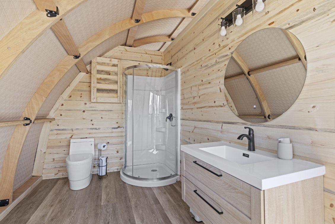 Alt text: "Interior of a stylish geodesic dome bathroom featuring wooden walls, a modern shower, toilet, and vanity with a round mirror. Ideal for luxury glamping or a geodome home setup."