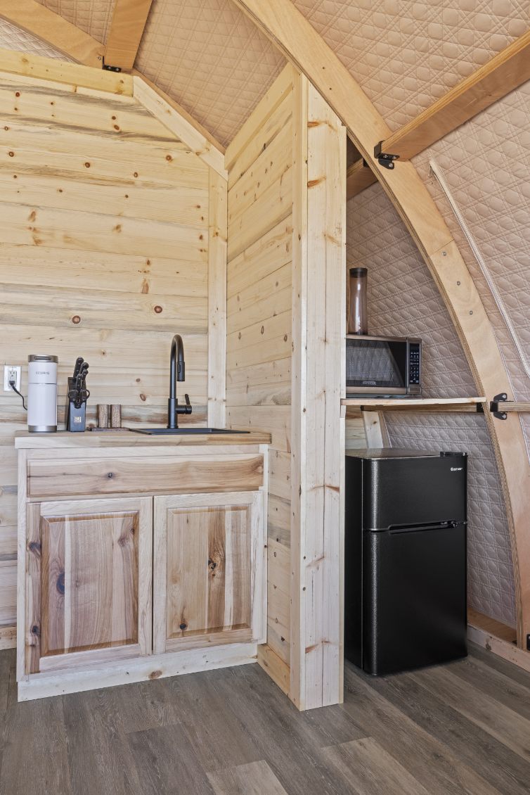 Alt text: "Interior view of a wooden glamping pod featuring a kitchenette with a sink, cabinets, and a small refrigerator. The stylish design highlights the use of natural wood and modern amenities, ideal for luxury camping or creating a cozy backyard office. Keywords: artemis pod, glamping pods for sale, geodesic dome kit."