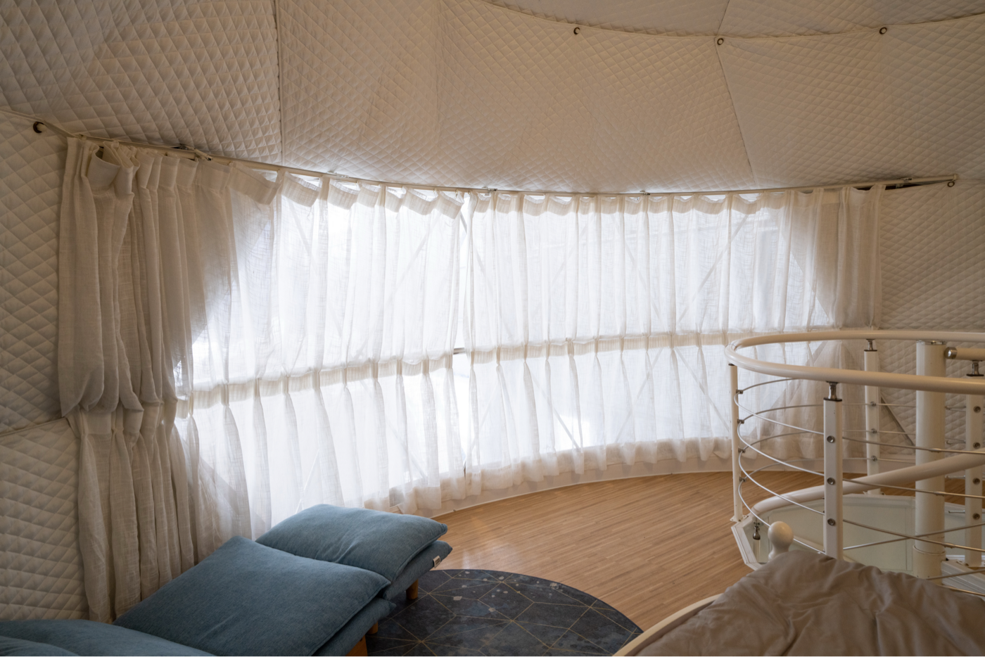 Interior view of a luxurious geodesic dome with quilted walls and large porthole windows, featuring a cozy seating area and spiral staircase. Ideal for glamping, sustainable living, or as a stylish backyard office. Explore premium geodome solutions for diverse needs.