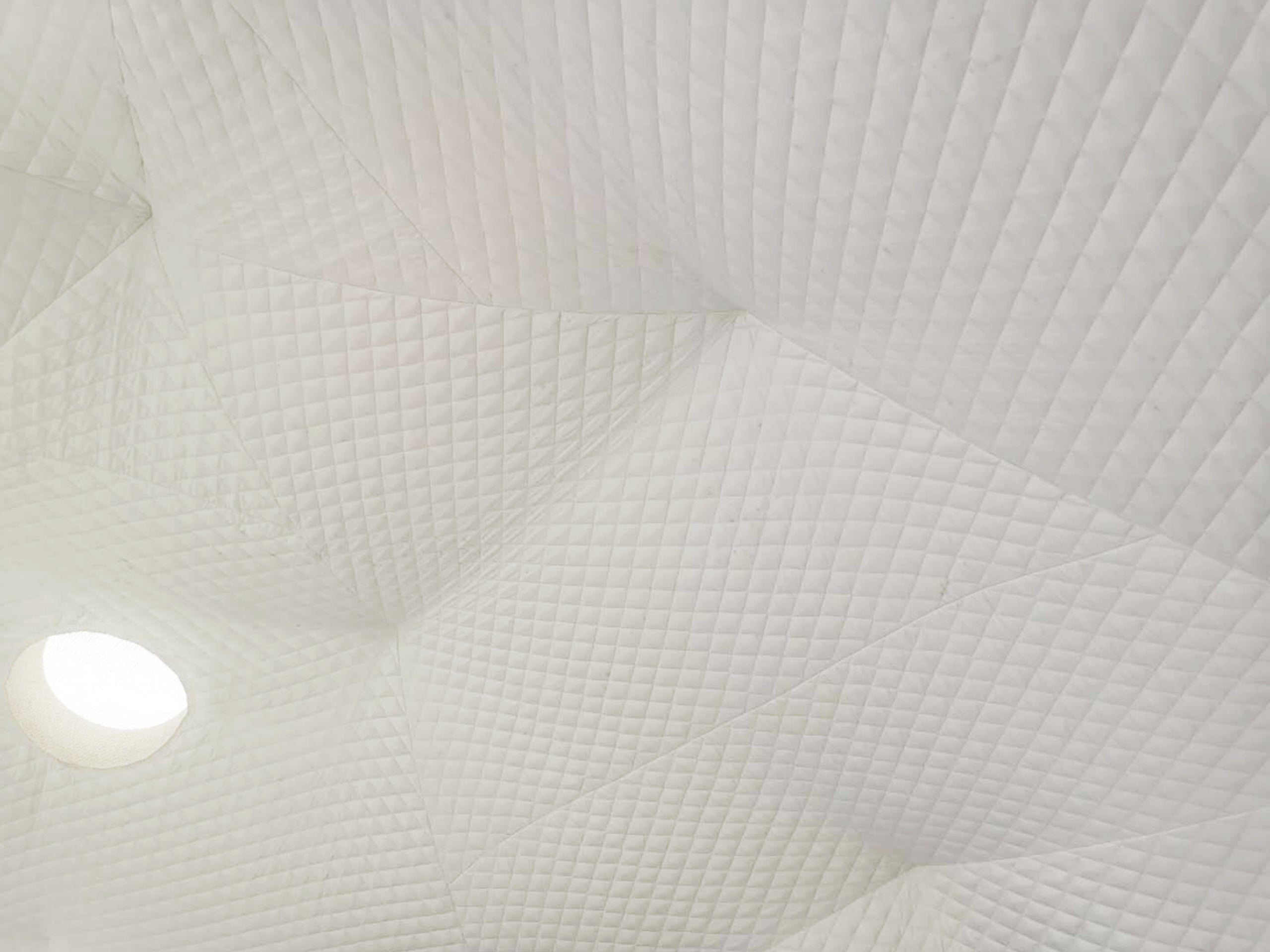 Alt text: "Interior view of a geodesic dome with quilted white fabric and a circular porthole window, showcasing the stylish and premium design of glamping solutions offered by the company, specializing in geodomes and dome homes."