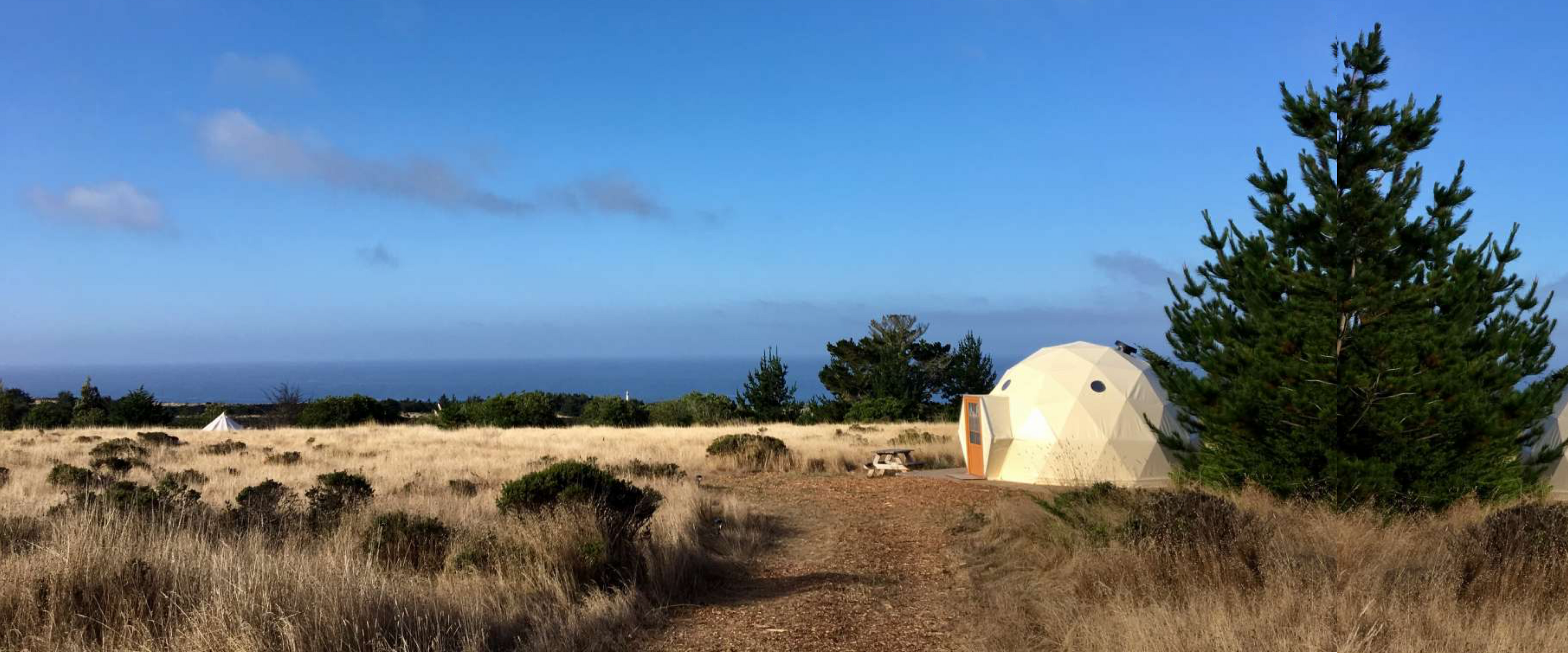 Alt text: "Geodesic dome with porthole windows set in a scenic outdoor landscape, ideal for glamping or as a luxury Airbnb. The dome is surrounded by natural greenery and overlooks a distant ocean view, showcasing the versatility of geodesic dome house kits for sustainable living and stylish accommodations."