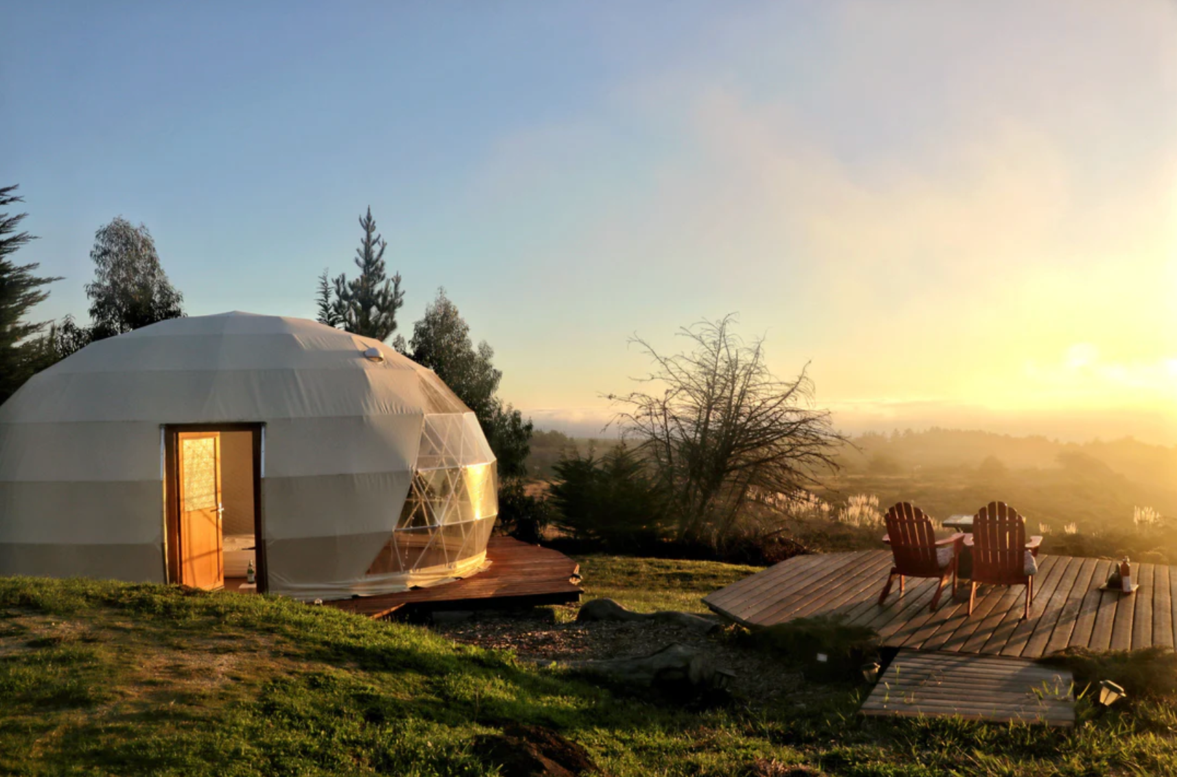 Alt text: "Scenic view of a geodesic dome house with porthole windows at sunset, surrounded by nature. The dome, ideal for glamping or as a backyard office, is set on a wooden deck with two chairs, offering a luxurious and sustainable living solution."