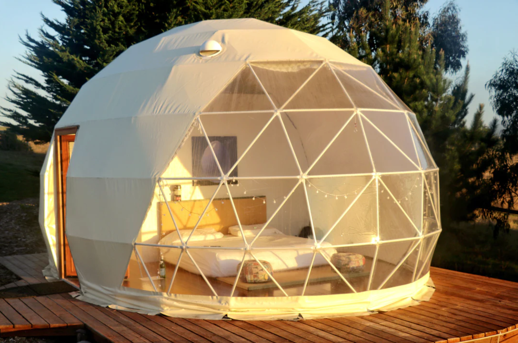 Alt text: "Luxury geodesic dome with porthole windows set up for glamping, featuring a cozy interior with a bed, located on a wooden deck surrounded by trees. Ideal for sustainable living, art studios, or as a stylish backyard dome. Available from a company specializing in high-quality glamping solutions."