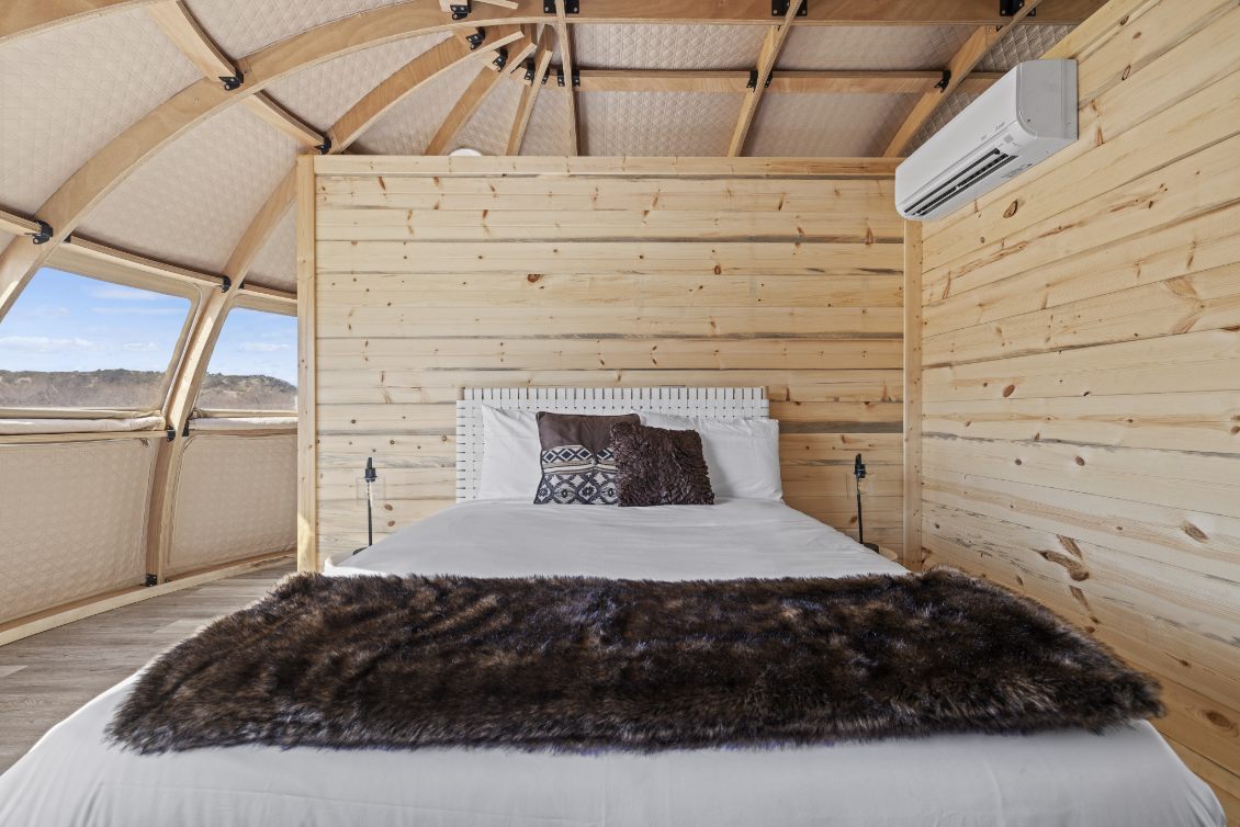 Alt text: &quot;Interior of a geodesic dome house featuring a cozy bedroom with wooden walls, a comfortable bed, and large porthole windows offering scenic views. Ideal for luxury glamping or a stylish backyard office. Keywords: geodesic dome, dome home, glamping pods.&quot;